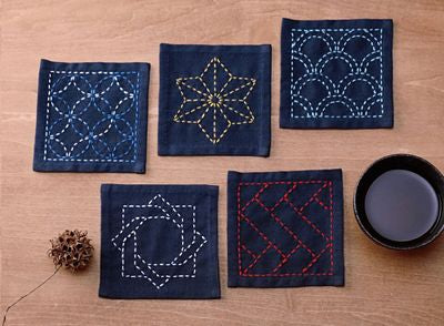 Five square dark indigo fabric coasters with colorful embroidered geometric patterns, arranged on a wooden tabletop next to a small black bowl and a pine cone.